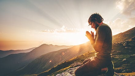 Man praying on a mountain top with the sunrise behind.